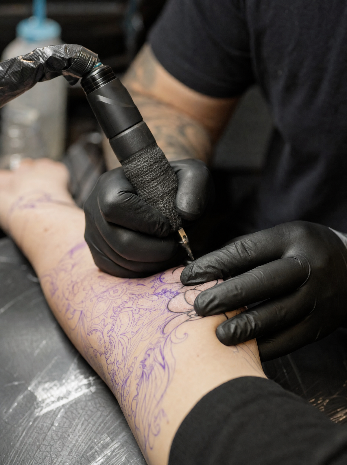 Black-gloved hands holding a tattoo machine over a stencilled forearm